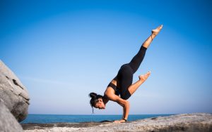 Woman performing a yoga pose on a rocky beach with ocean view and clear blue sky.